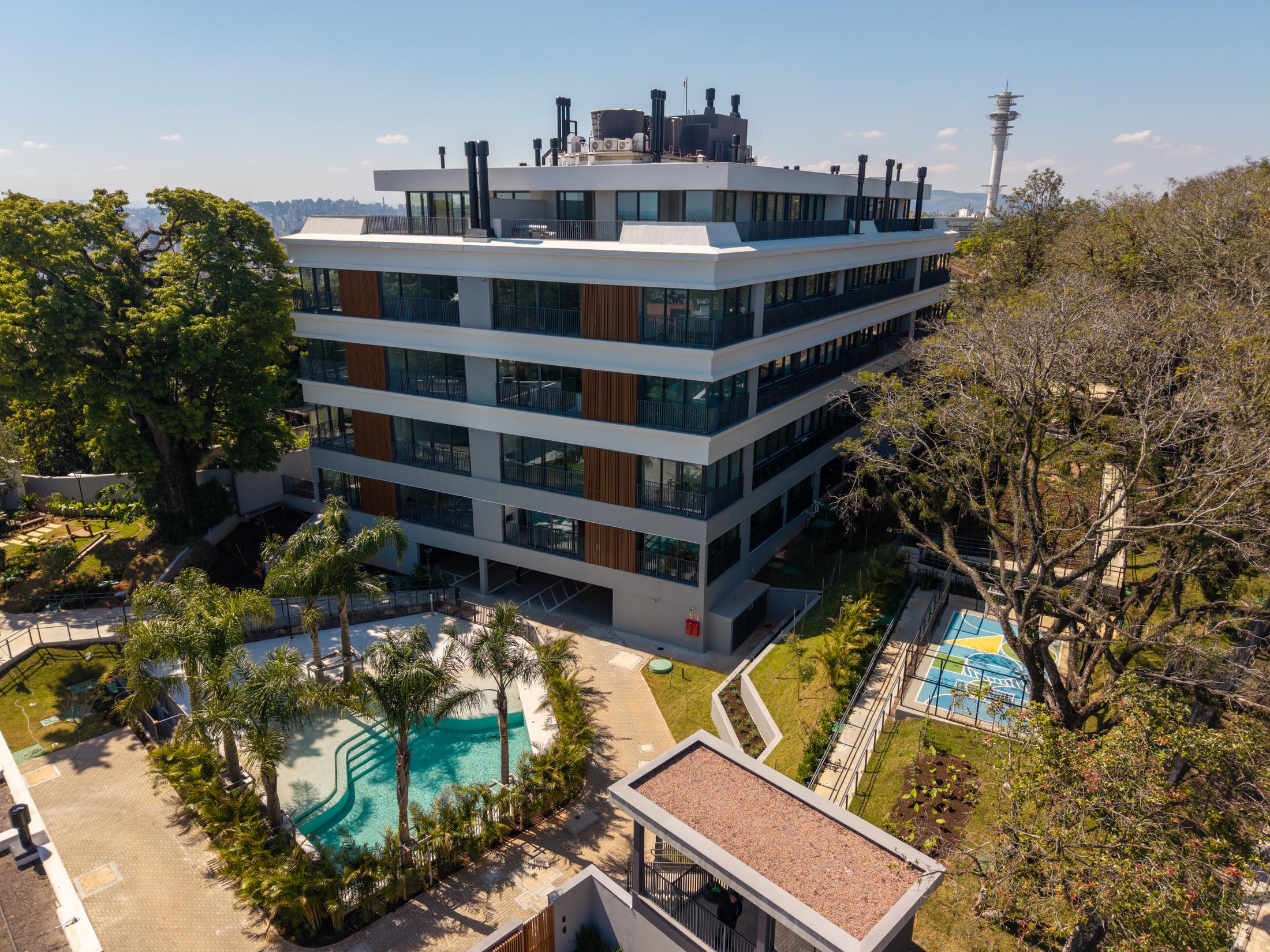 Foto aérea do Aria pronto: o prédio com fachada de madeira, a piscina entre palmeiras, a quadra e as árvores maduras ao redor