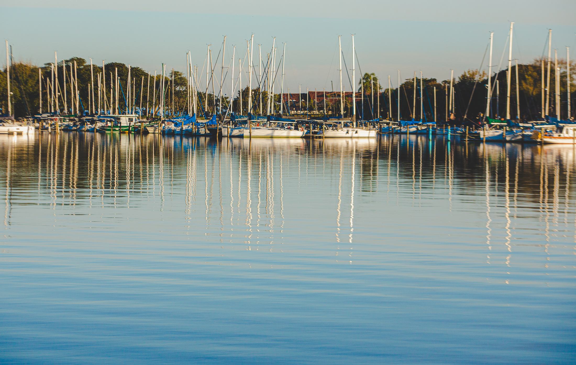 Marina no Guaíba em um dia de sol, com veleiros e o céu azul refletidos na água calma