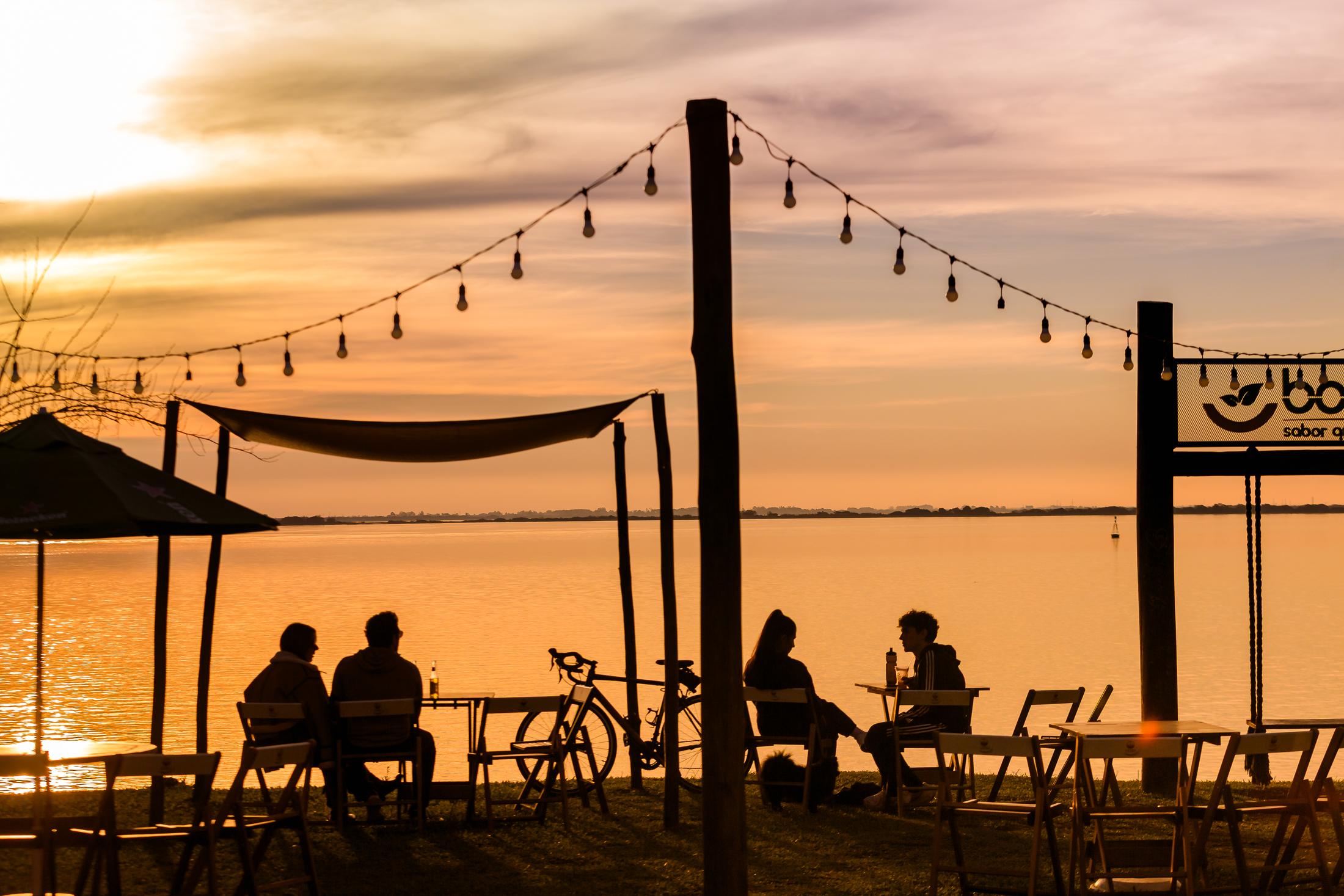 Pessoas à mesa em um bar na orla do Guaíba ao pôr do sol