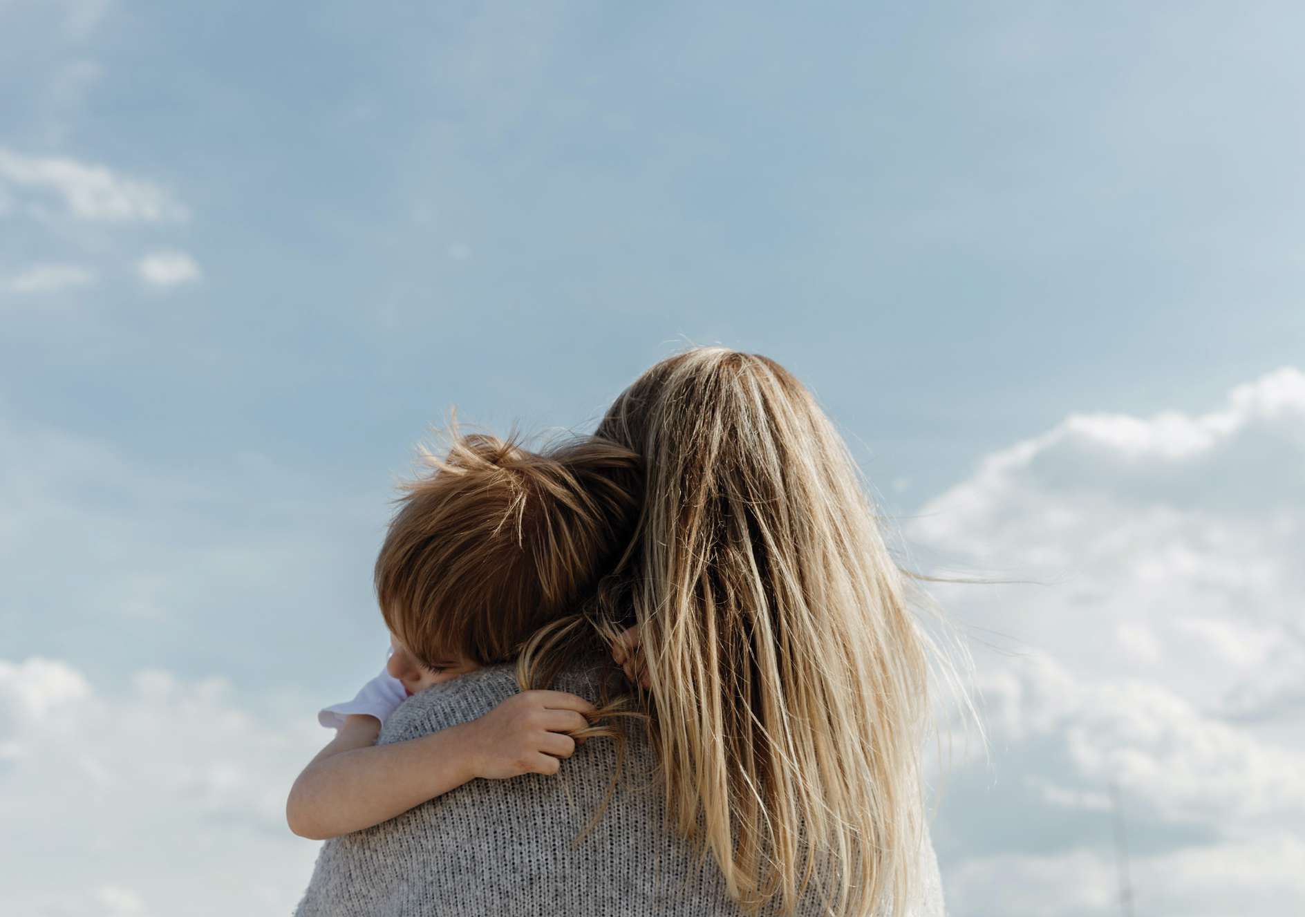 Mãe abraçando o filho no colo, de costas, diante de um céu azul com nuvens